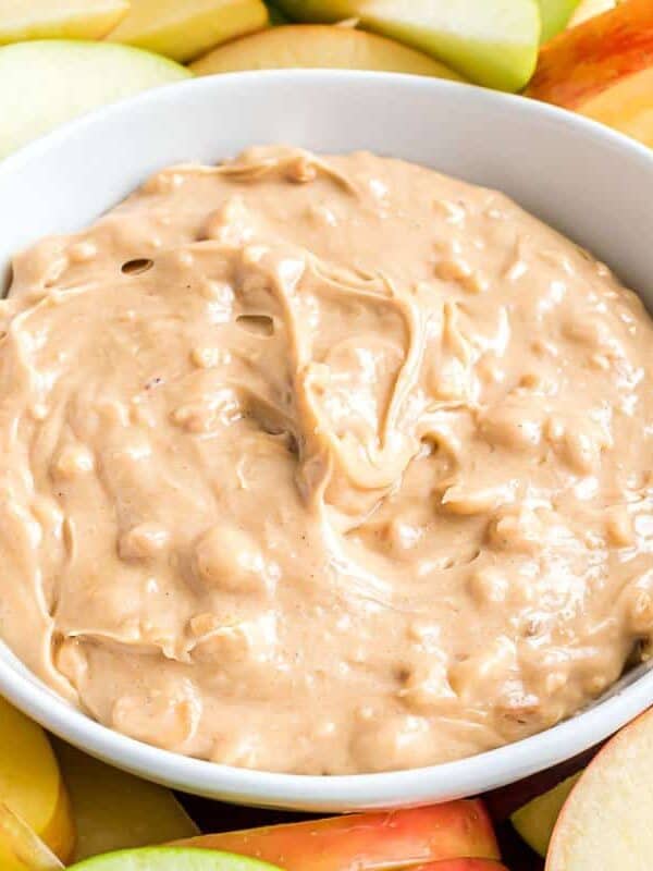 A white bowl filled with creamy peanut butter dip surrounded by sliced green and red apples.