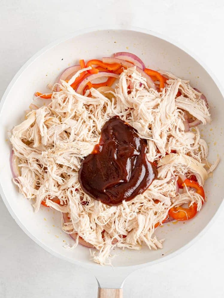 Shredded chicken, sliced red bell peppers, and onions in a white skillet topped with barbecue sauce.