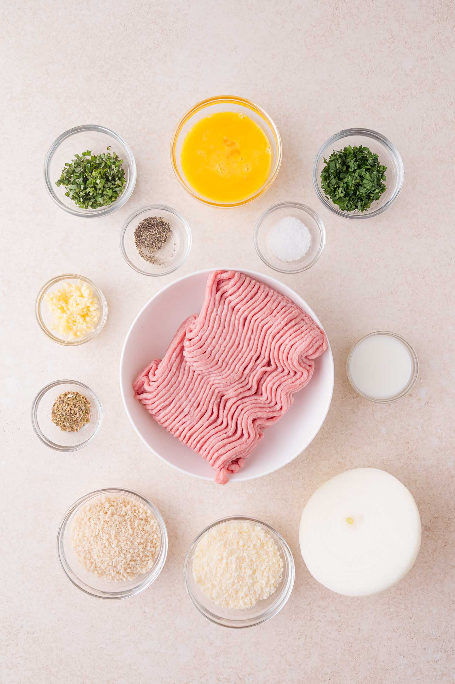 A bowl of raw ground meat surrounded by small bowls of chopped herbs, beaten egg, spices, minced garlic, bread crumbs, grated cheese, milk, and a halved onion on a light surface.