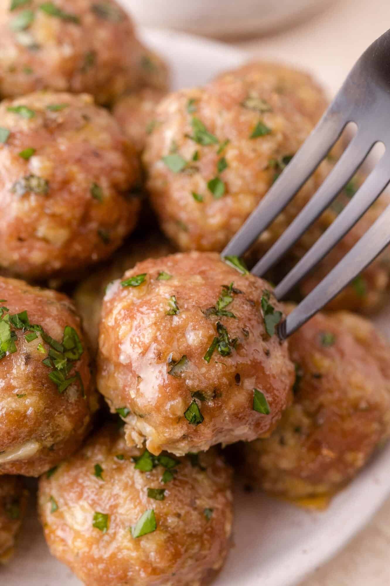 A close-up of a fork picking up a seasoned meatball garnished with chopped herbs from a plate of several meatballs.