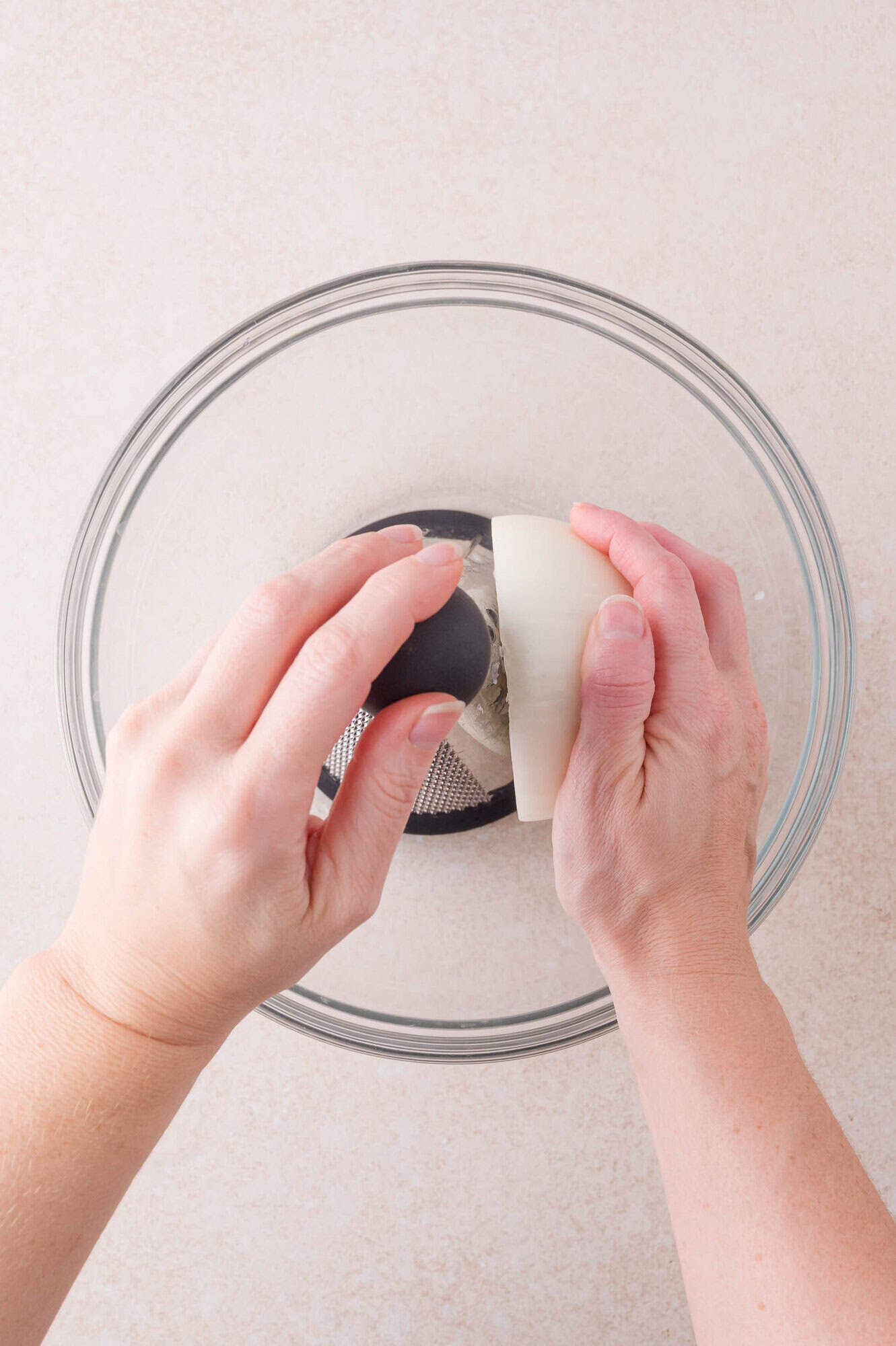 A person grates a white onion over a clear glass bowl using a metal grater.