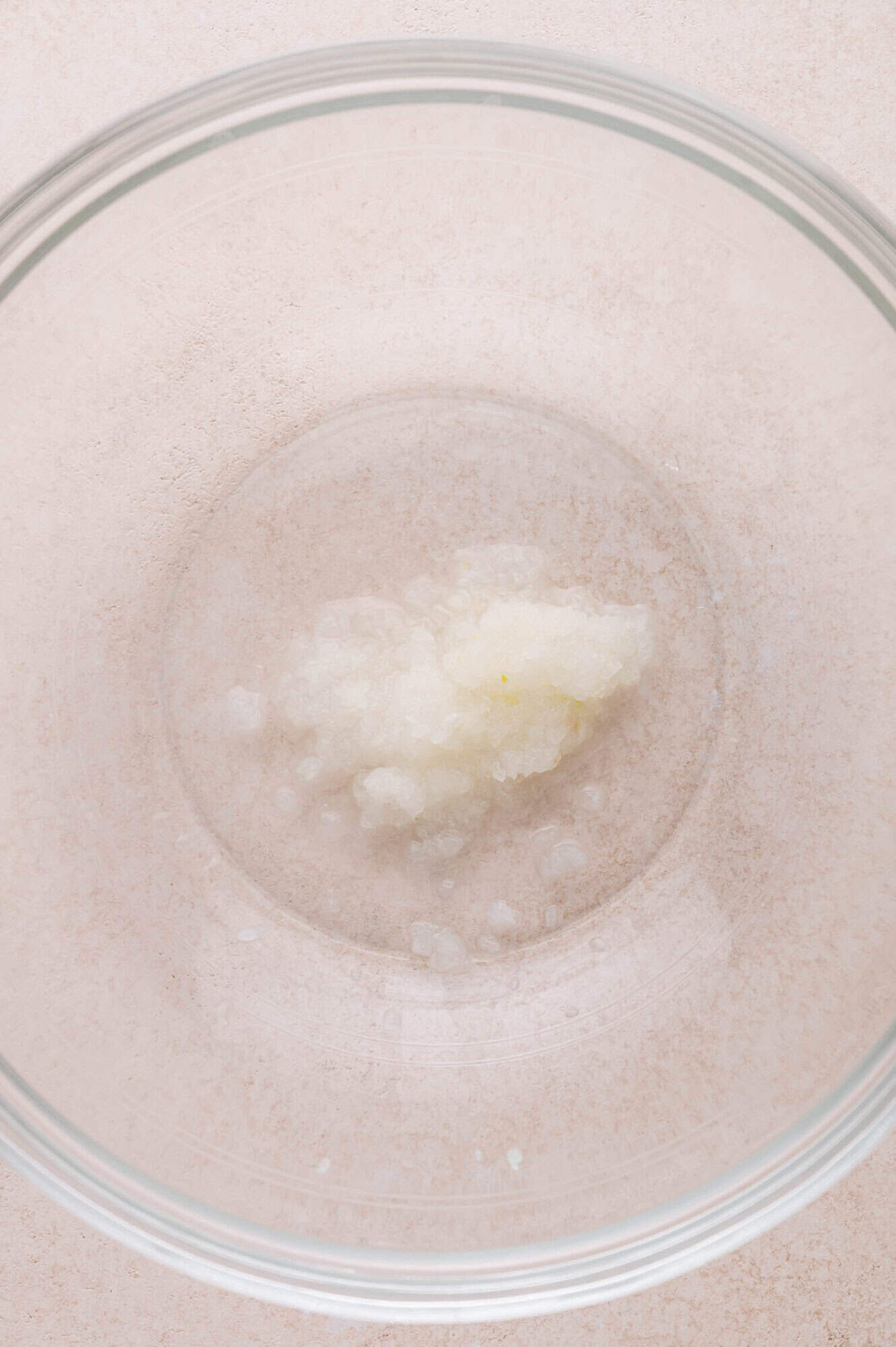 A small amount of grated onion in the center of a large, clear glass mixing bowl on a light-colored surface.