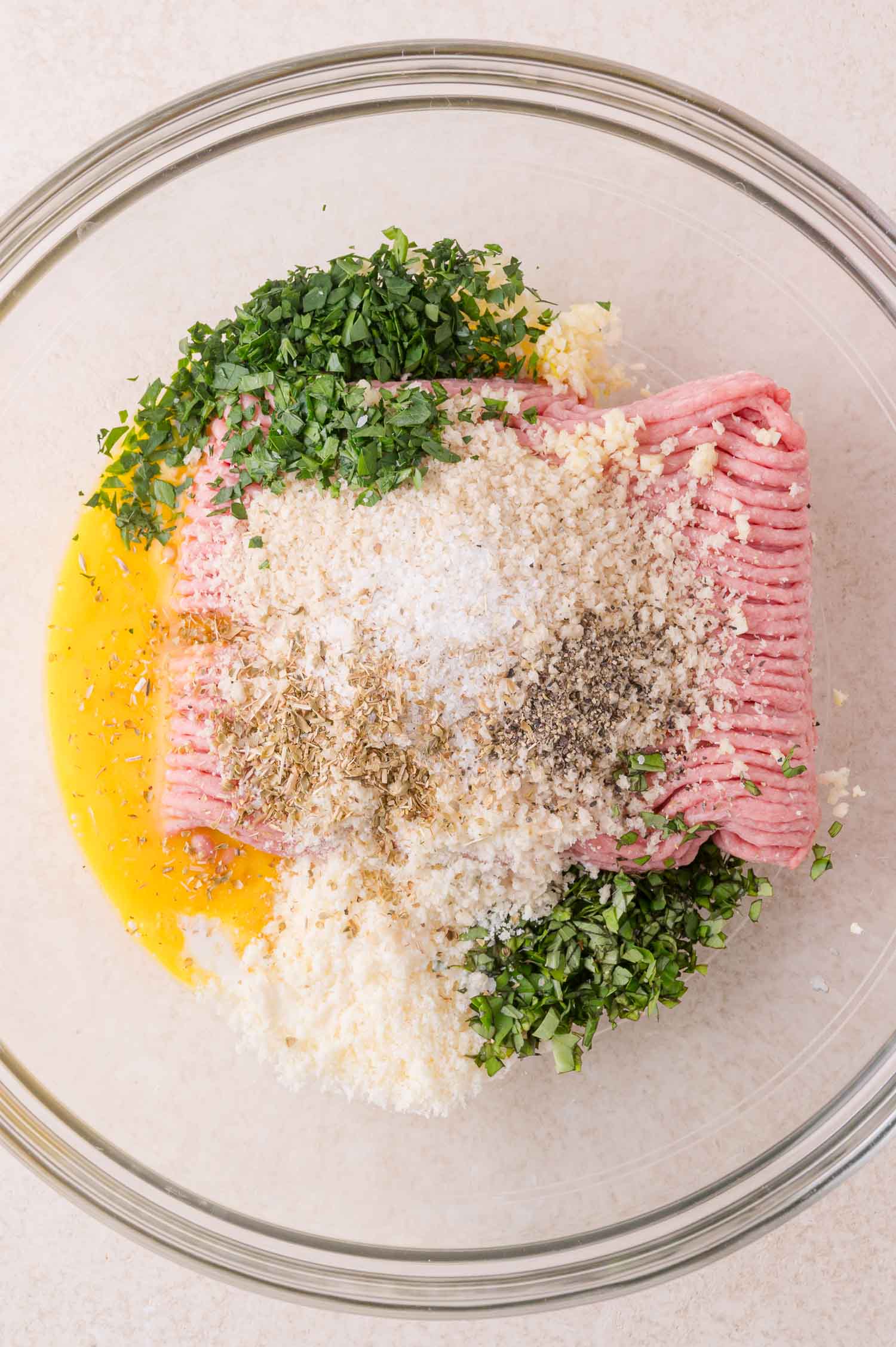 A glass bowl containing ground meat, chopped herbs, grated cheese, panko breadcrumbs, an egg, and seasonings, ready to be mixed.