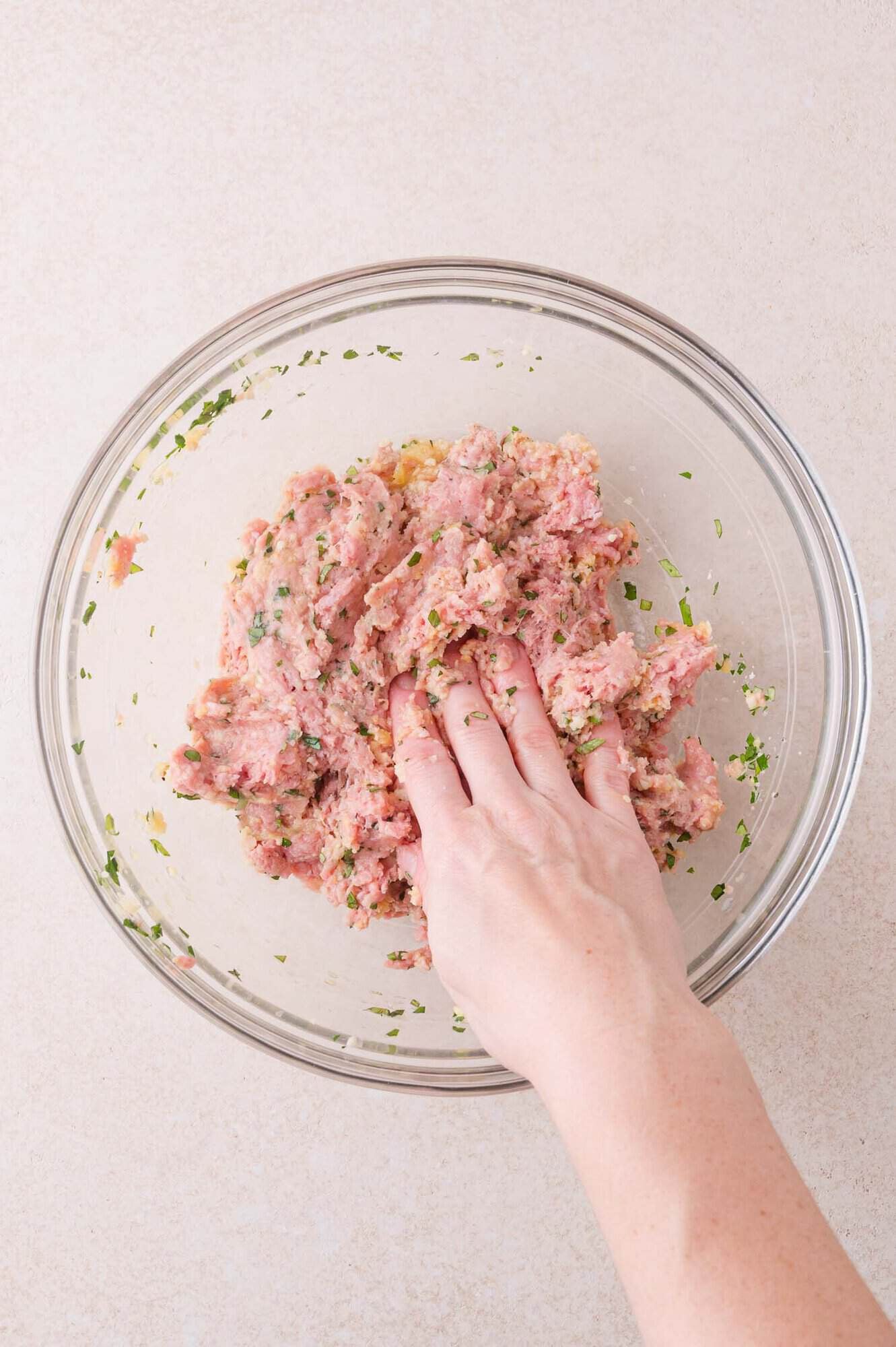 A hand mixes ground meat and herbs in a glass bowl on a light-colored surface.