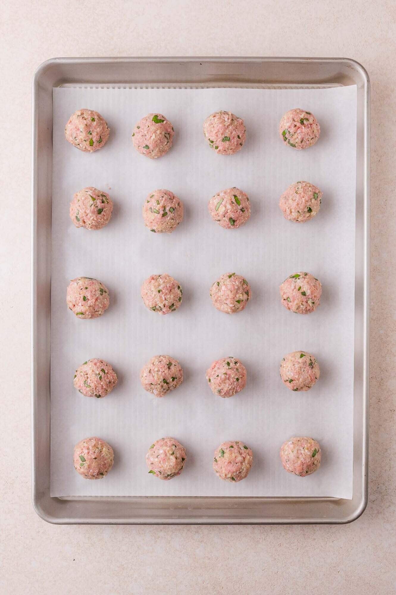 A baking sheet lined with parchment paper holds twenty evenly spaced, uncooked meatballs with visible herbs mixed in.