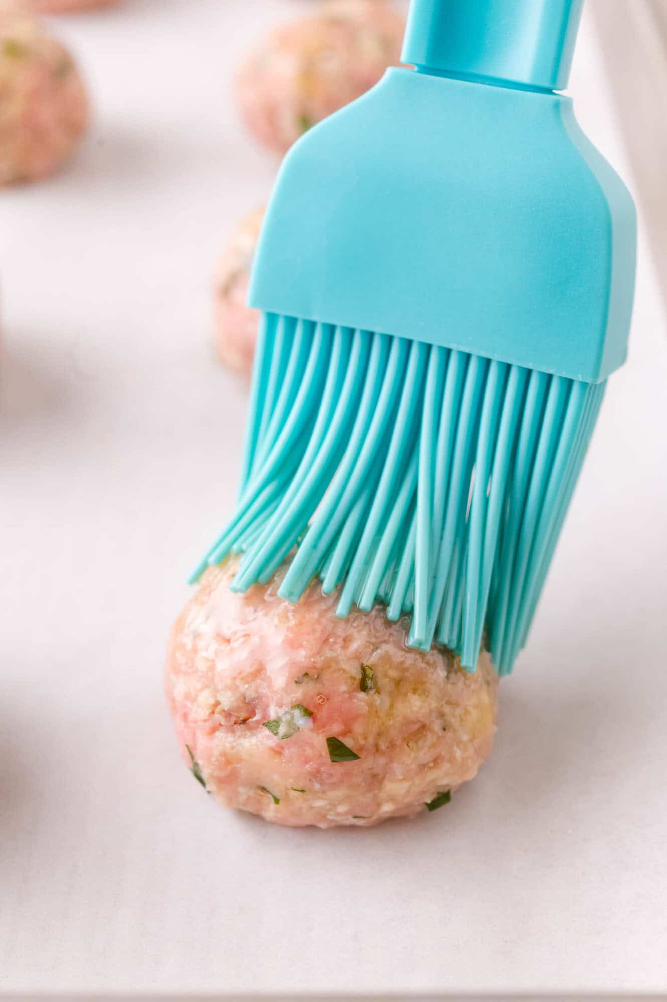A blue silicone brush applying oil to a raw meatball on a parchment-lined baking sheet.