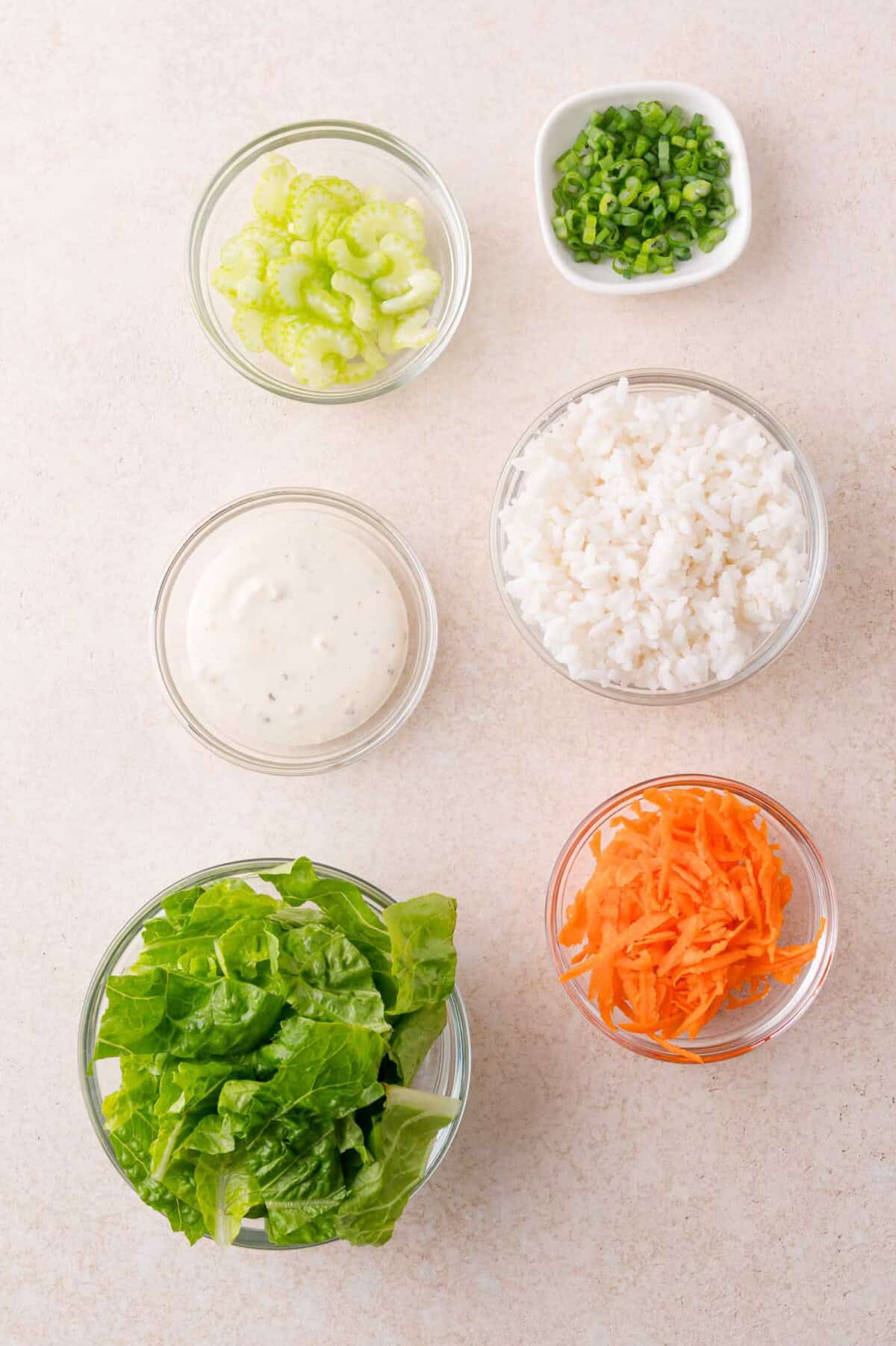 Six bowls with chopped celery, sliced green onions, cooked white rice, ranch dressing, shredded lettuce, and grated carrots on a light surface.