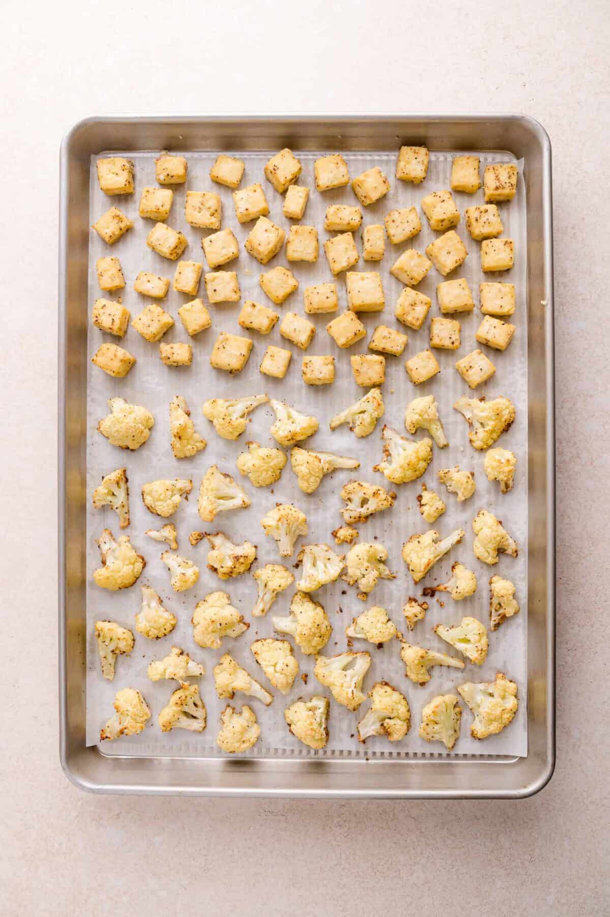 Baking sheet lined with parchment paper holding evenly spaced roasted cauliflower florets on the bottom half and roasted tofu cubes on the top half.