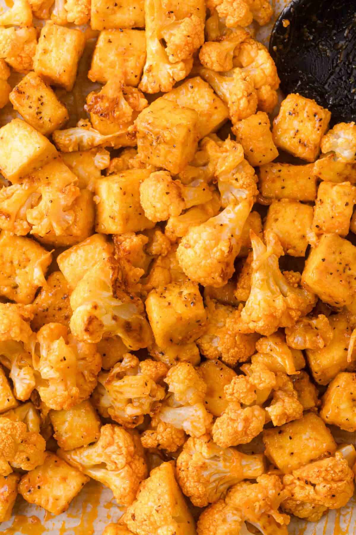 Roasted cauliflower florets and tofu cubes coated in spices, arranged on a baking sheet with a black spatula partially visible.