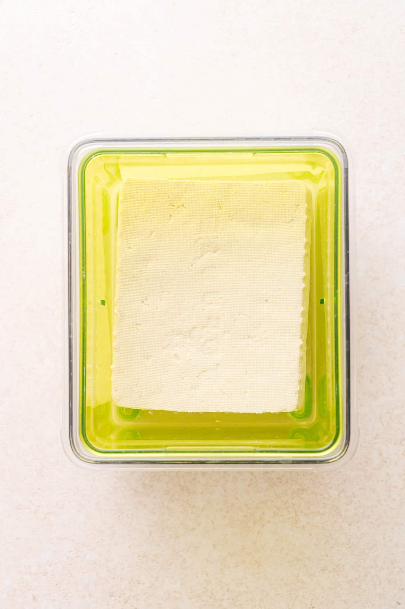 A block of tofu sits in a transparent container with a green base, filled with liquid, viewed from above on a light surface.