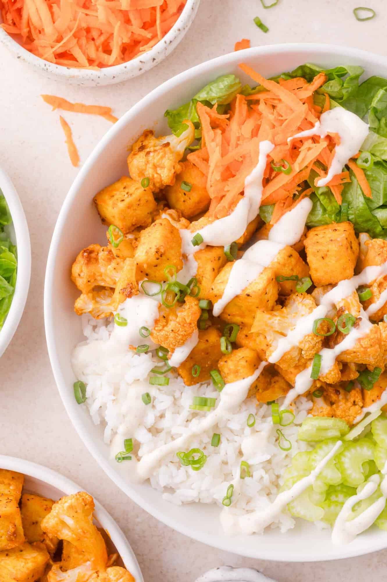 A bowl filled with white rice, lettuce, shredded carrots, seasoned tofu and cauliflower, and drizzled with a ranch dressing, garnished with chopped green onions.