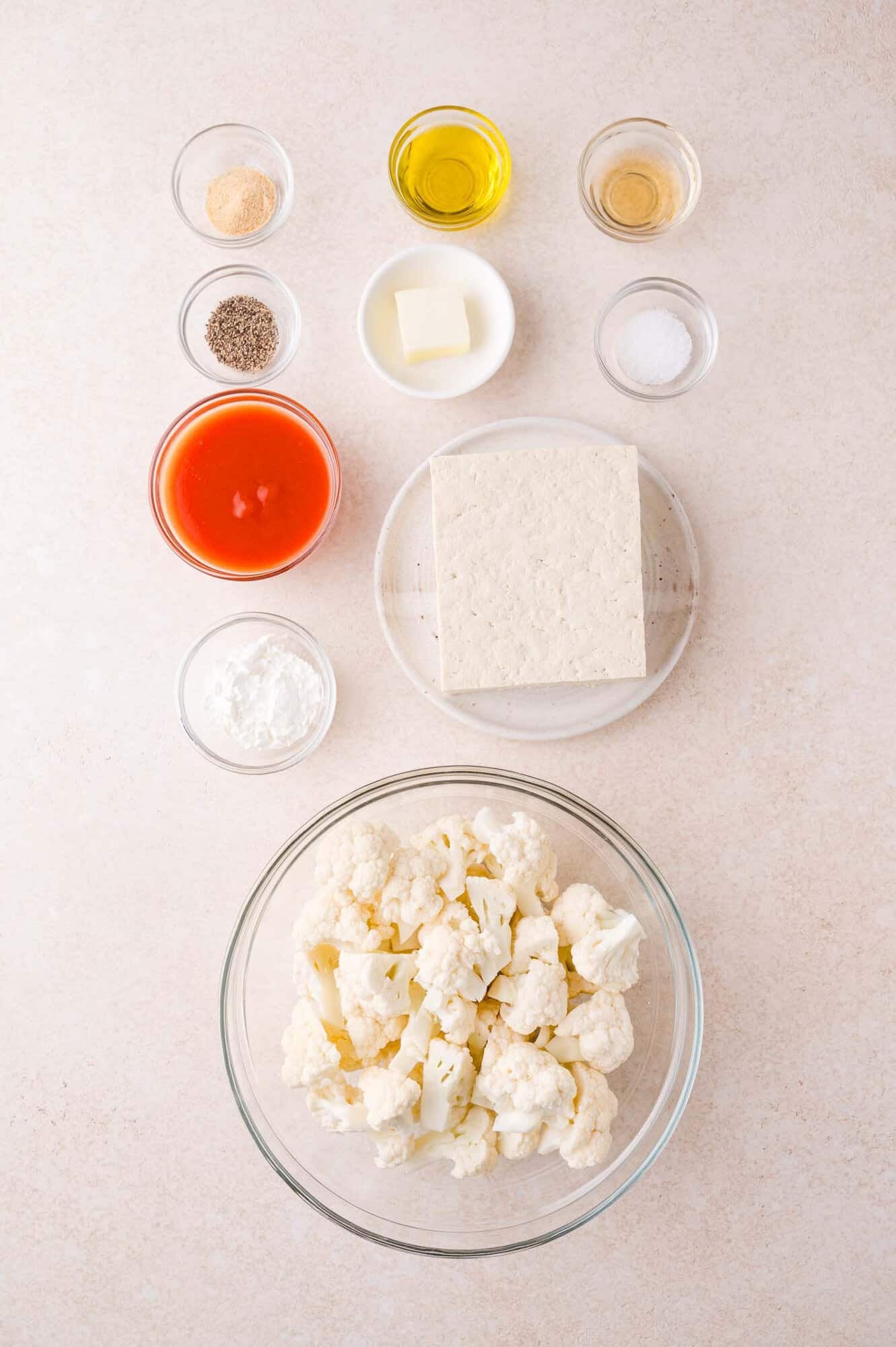 A glass bowl of cauliflower florets, a block of tofu, and small bowls containing hot sauce, oil, butter, vinegar, cornstarch, garlic powder, pepper, and salt arranged on a light surface.