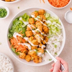 A bowl with cubed seasoned tofu, shredded lettuce, sliced cucumber, shredded carrots, white rice, and a drizzle of ranch dressing, with a hand holding a fork.