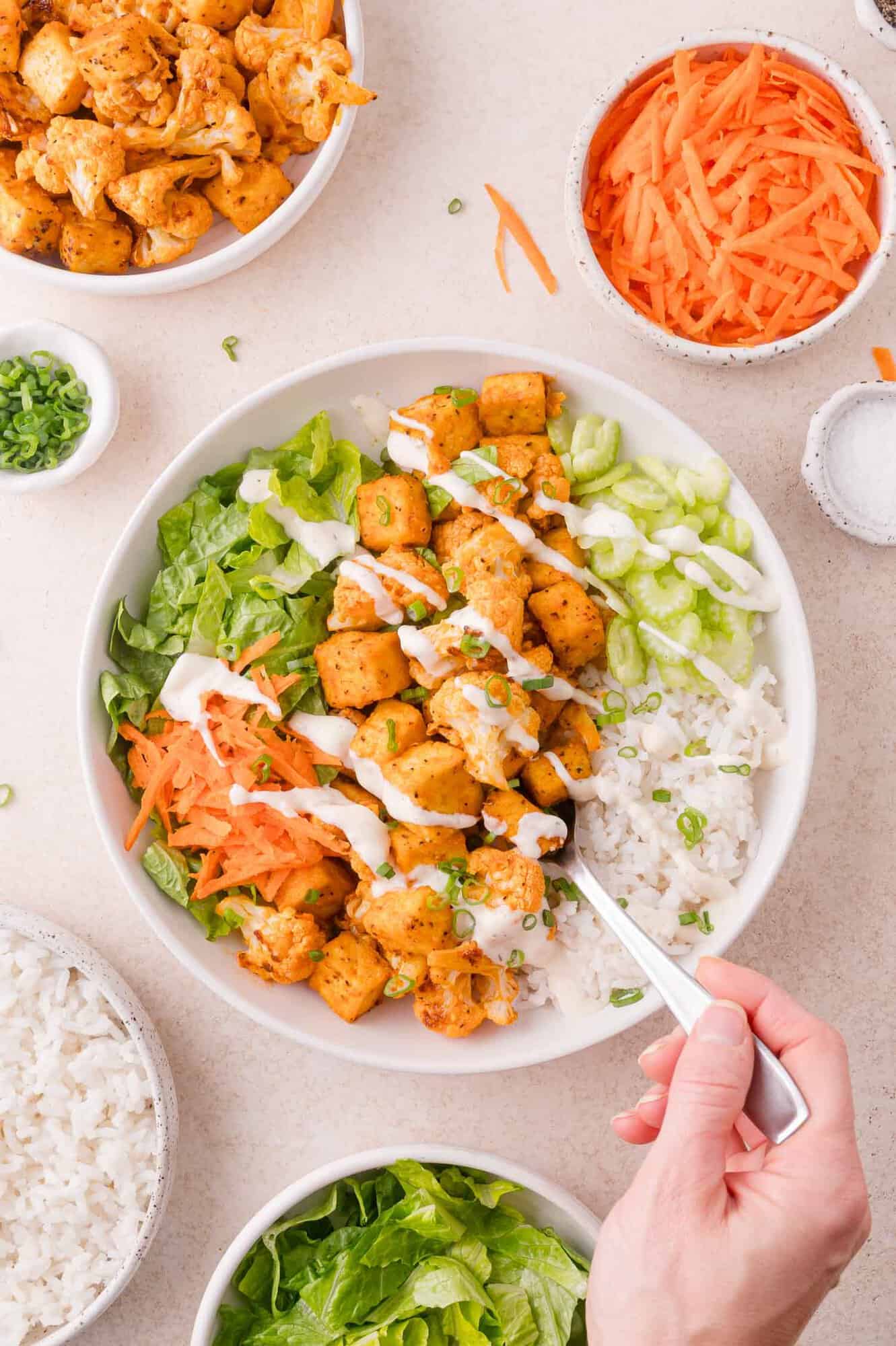 A bowl of rice, lettuce, shredded carrots, seasoned tofu pieces, and ranch dressing, with a hand holding a fork. Surrounding bowls contain tofu, carrots, rice, and lettuce.