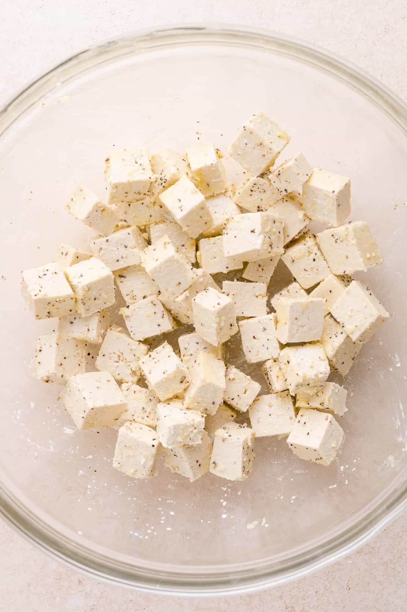 Cubed tofu seasoned with black pepper and spices in a clear glass bowl on a light-colored surface.