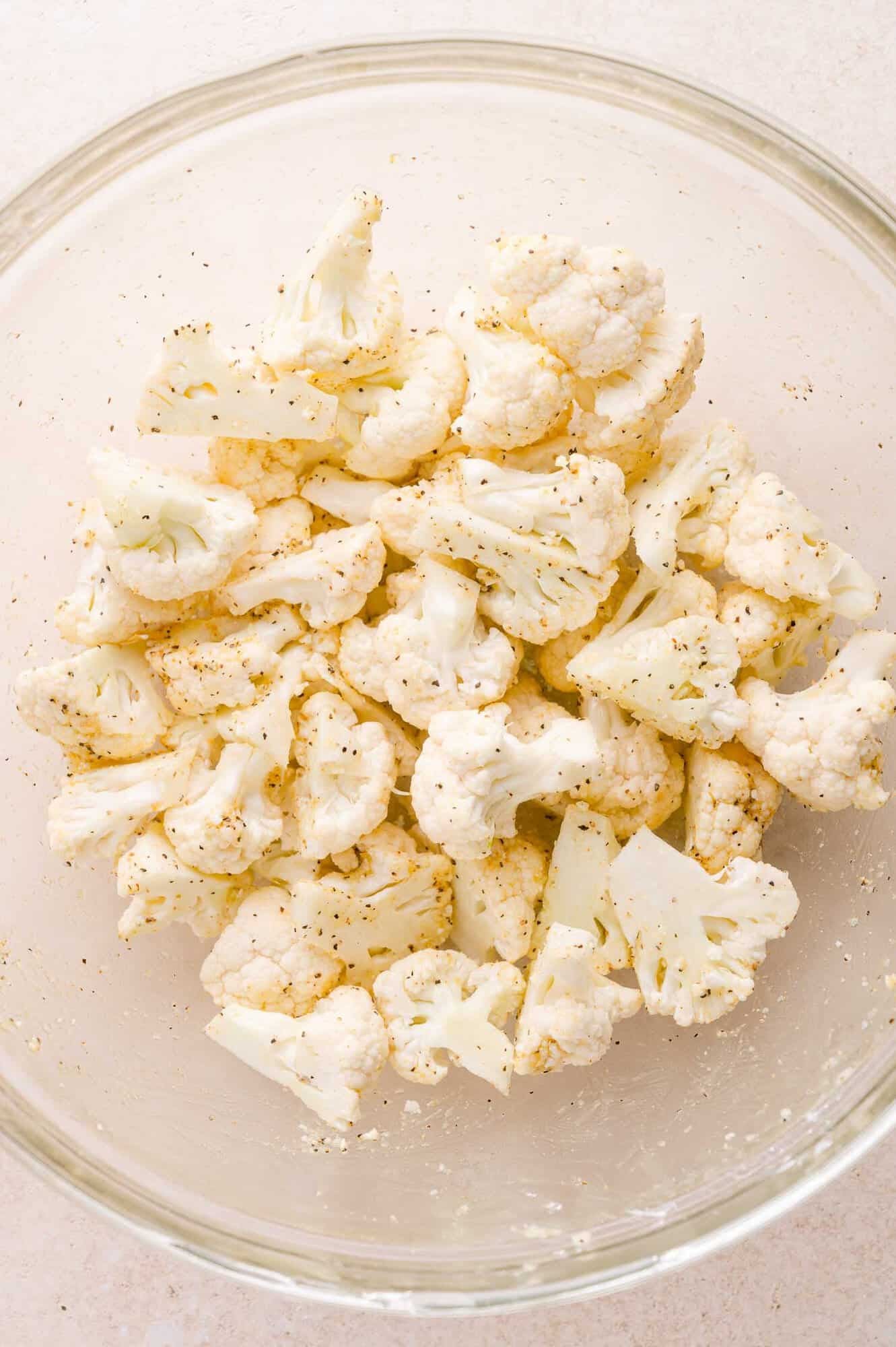 Raw cauliflower florets in a glass bowl, seasoned with salt, pepper, and other spices.