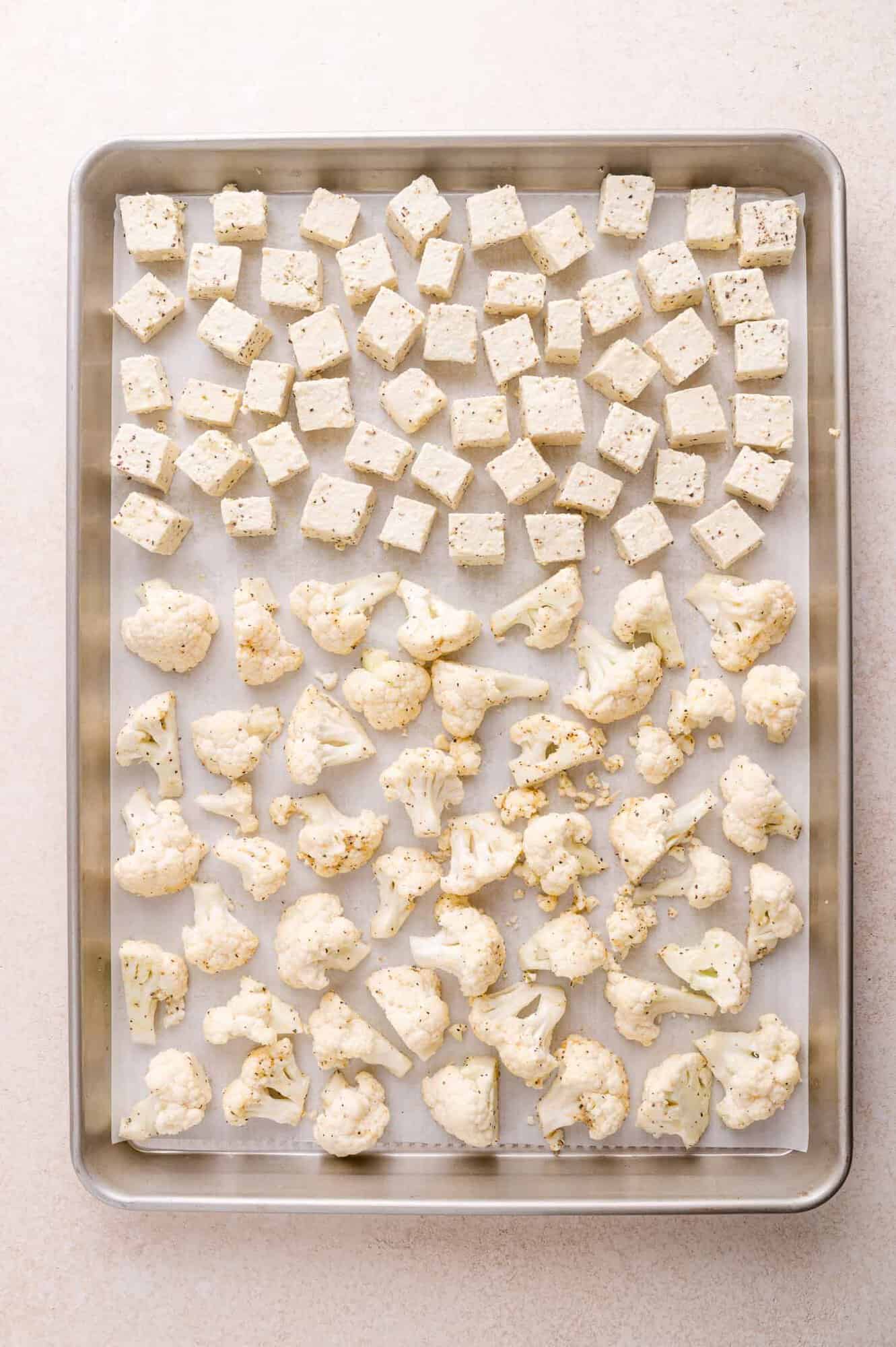 A baking sheet with evenly spaced cubes of tofu and cauliflower florets, both seasoned, on parchment paper.