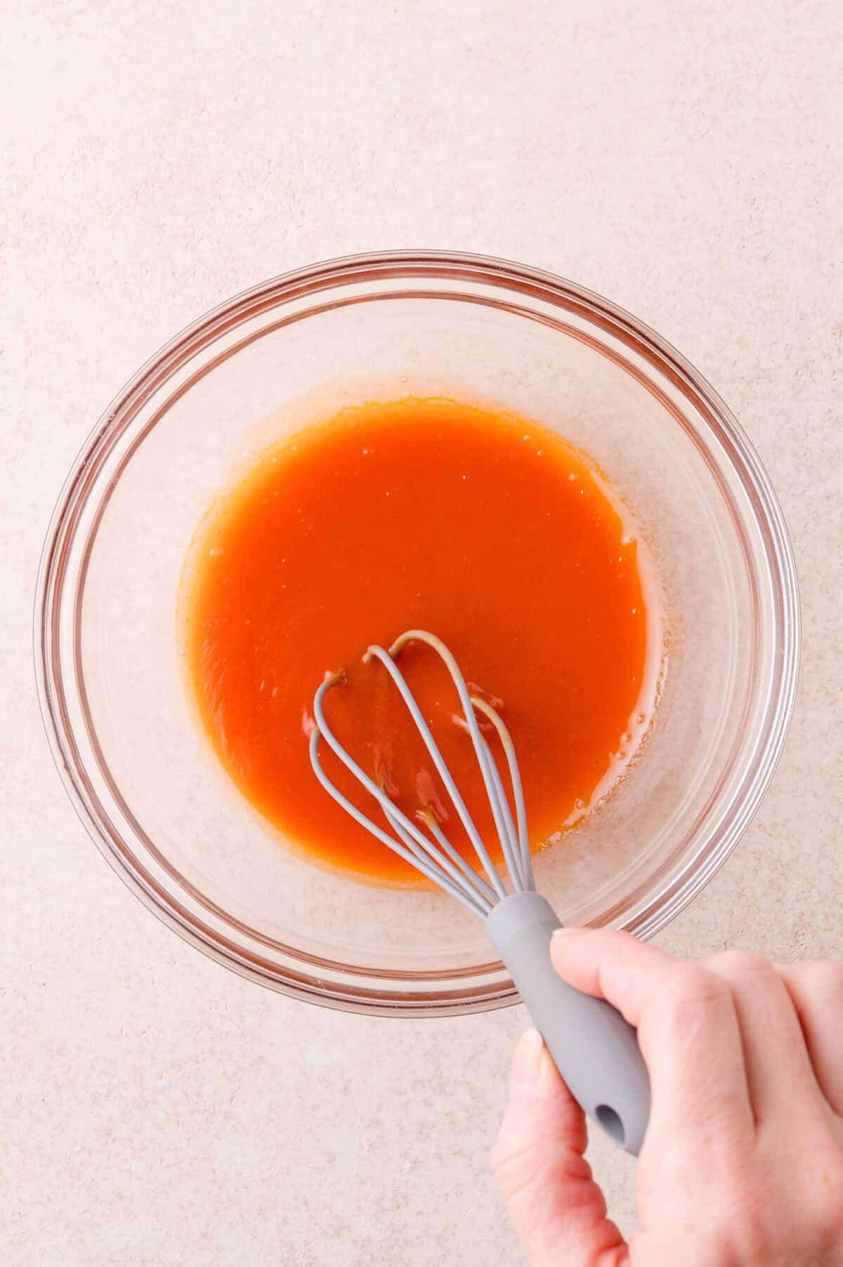 A hand holding a whisk mixes an orange liquid in a clear glass bowl on a light-colored surface.