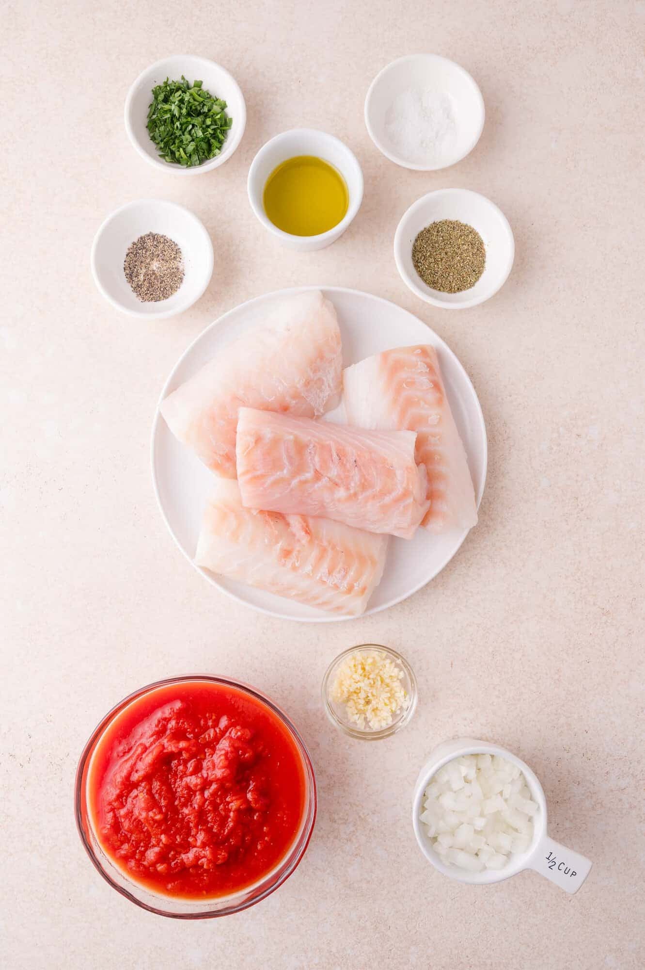 Raw cod fillets on a plate, surrounded by bowls of chopped herbs, olive oil, salt, pepper, Italian seasoning, minced garlic, chopped onion, and crushed tomatoes.