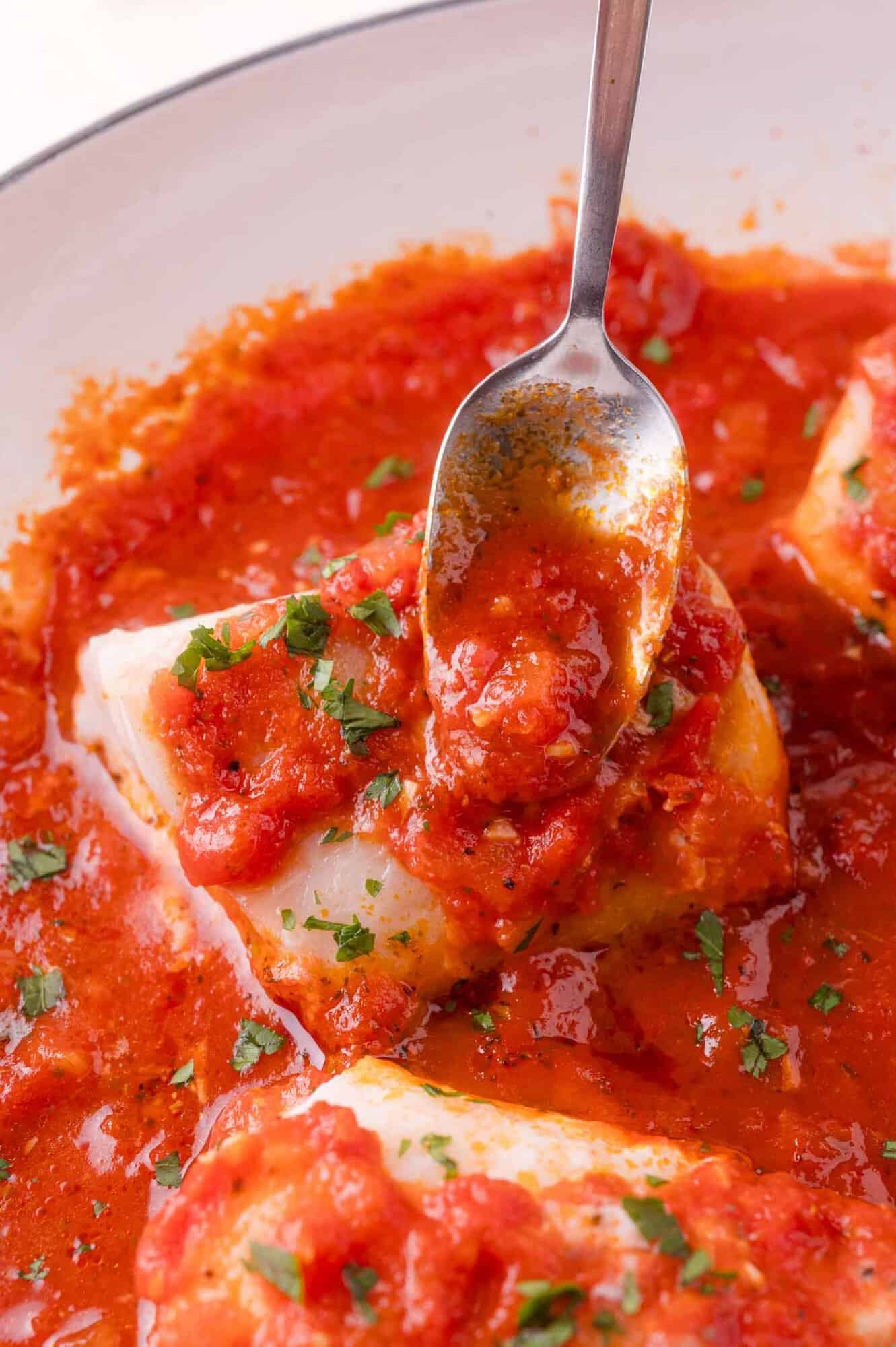 A spoon pours tomato sauce over a piece of white fish, garnished with chopped herbs, in a white dish.