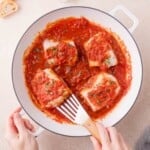 Four pieces of cod are cooked in a skillet with chunky tomato sauce and garnished with herbs; a spatula is lifting one piece.