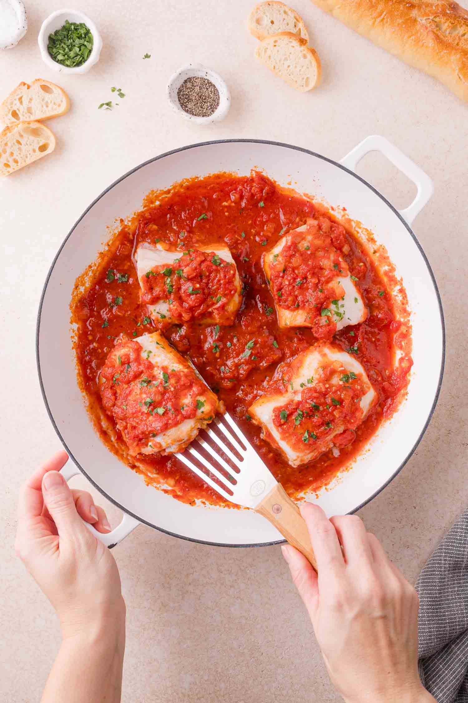 A person serves white cod fillets in tomato sauce from a white pan, surrounded by small bowls of herbs, pepper, and slices of baguette.
