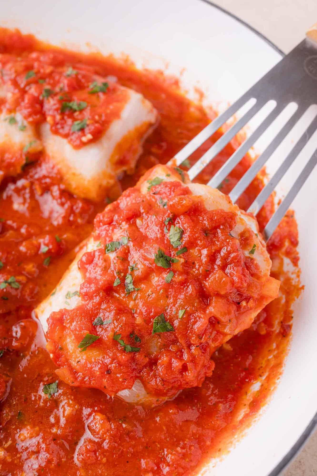 A piece of white fish topped with tomato sauce and herbs is lifted from a pan filled with tomato sauce using a metal spatula.