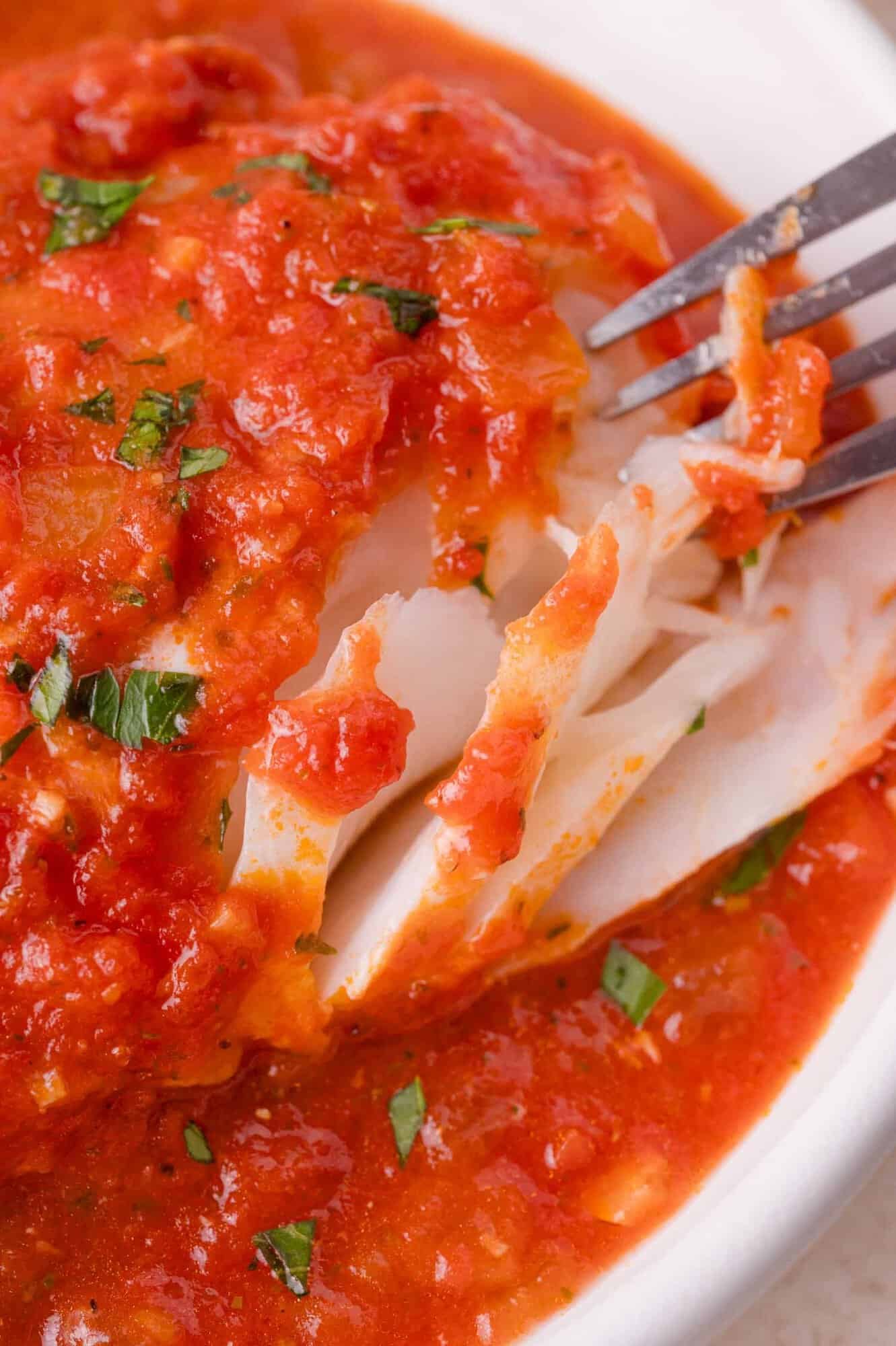 A close-up of a flaky cod fillet topped with chunky tomato sauce and garnished with chopped herbs, being pulled apart with a fork.