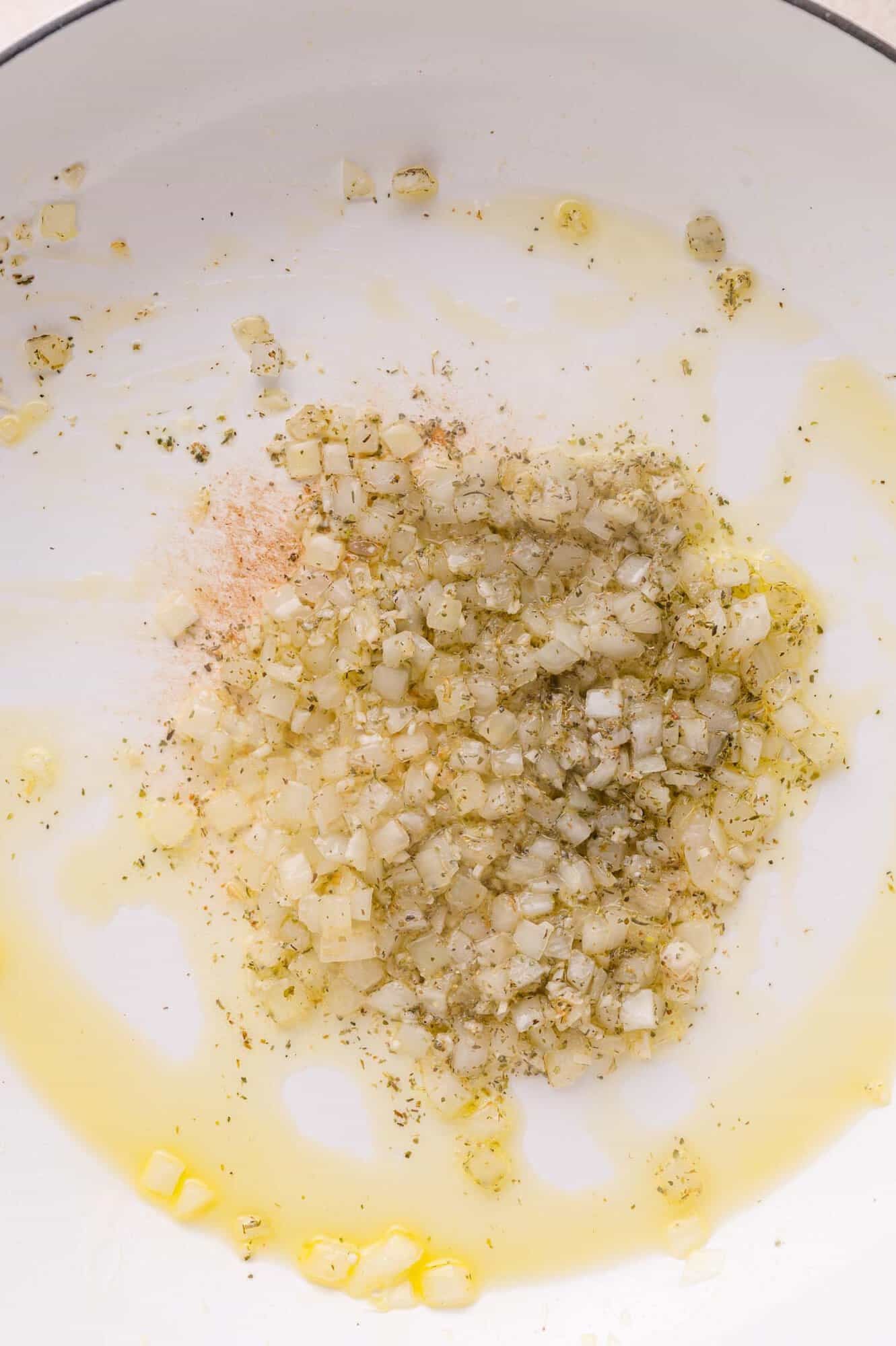 Diced onions sautéing in oil with herbs and spices in a white pan.