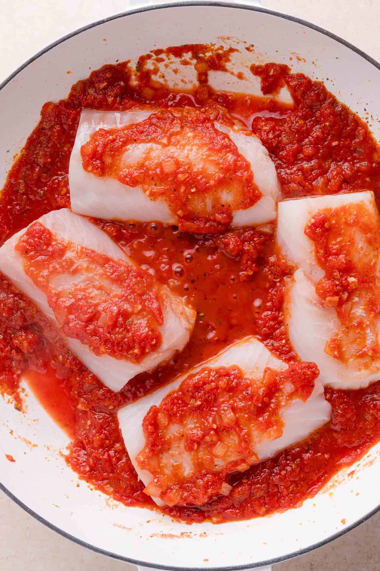 Four pieces of raw white cod in a pan, topped with chunky tomato sauce, ready to be cooked.