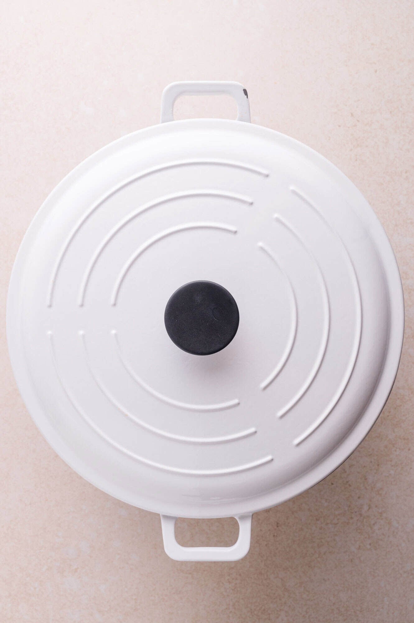 A white enameled cast iron Dutch oven with a black knob sits on a light-colored surface, viewed from above.