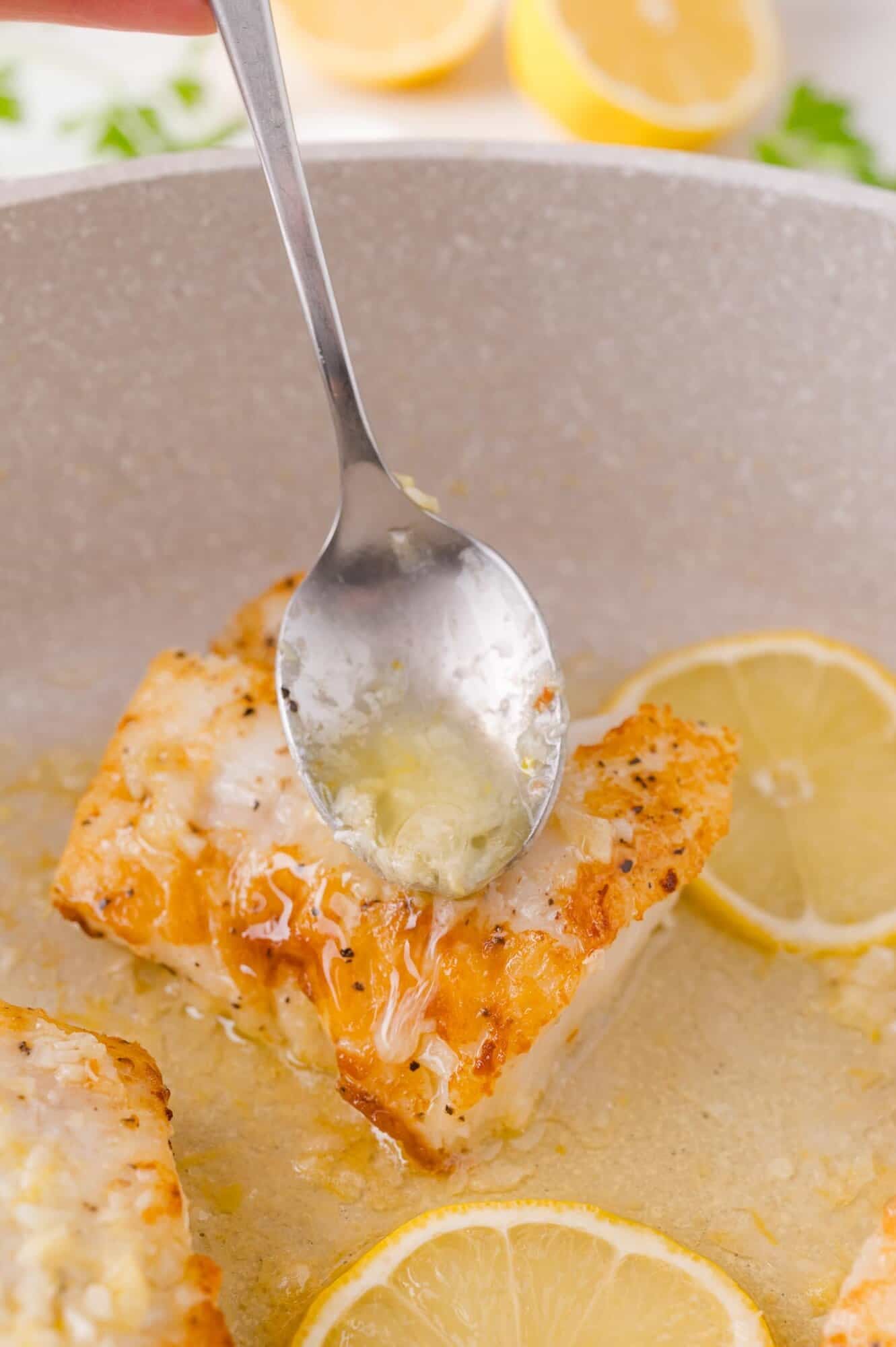 A spoon drizzles melted garlic butter over a piece of seared fish in a pan, surrounded by lemon slices.