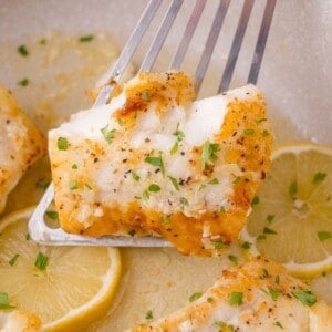 A piece of seasoned, cooked cod is lifted by a metal spatula above lemon slices, garnished with chopped herbs.