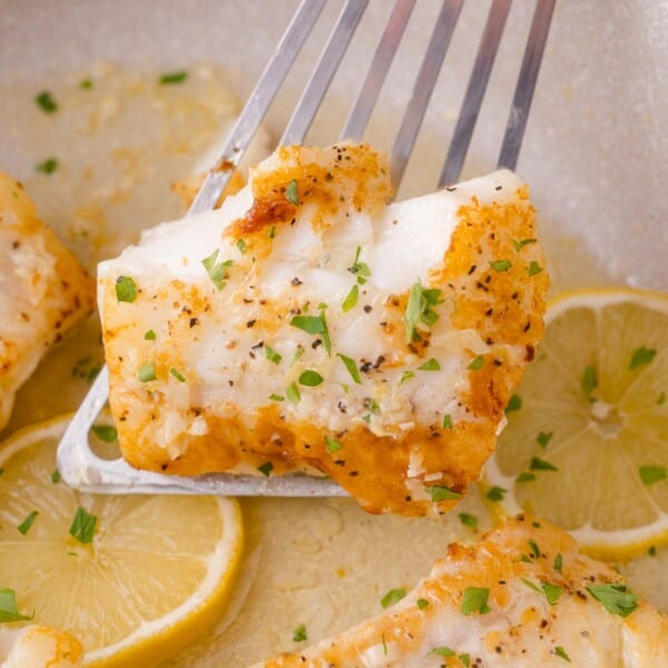 A piece of seasoned, cooked cod is lifted by a metal spatula above lemon slices, garnished with chopped herbs.