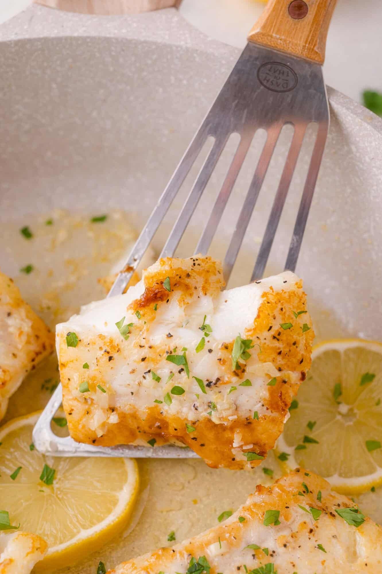 A metal spatula lifts a seasoned, cooked white lemon butter cod fillet garnished with chopped herbs, with lemon slices visible in the background.