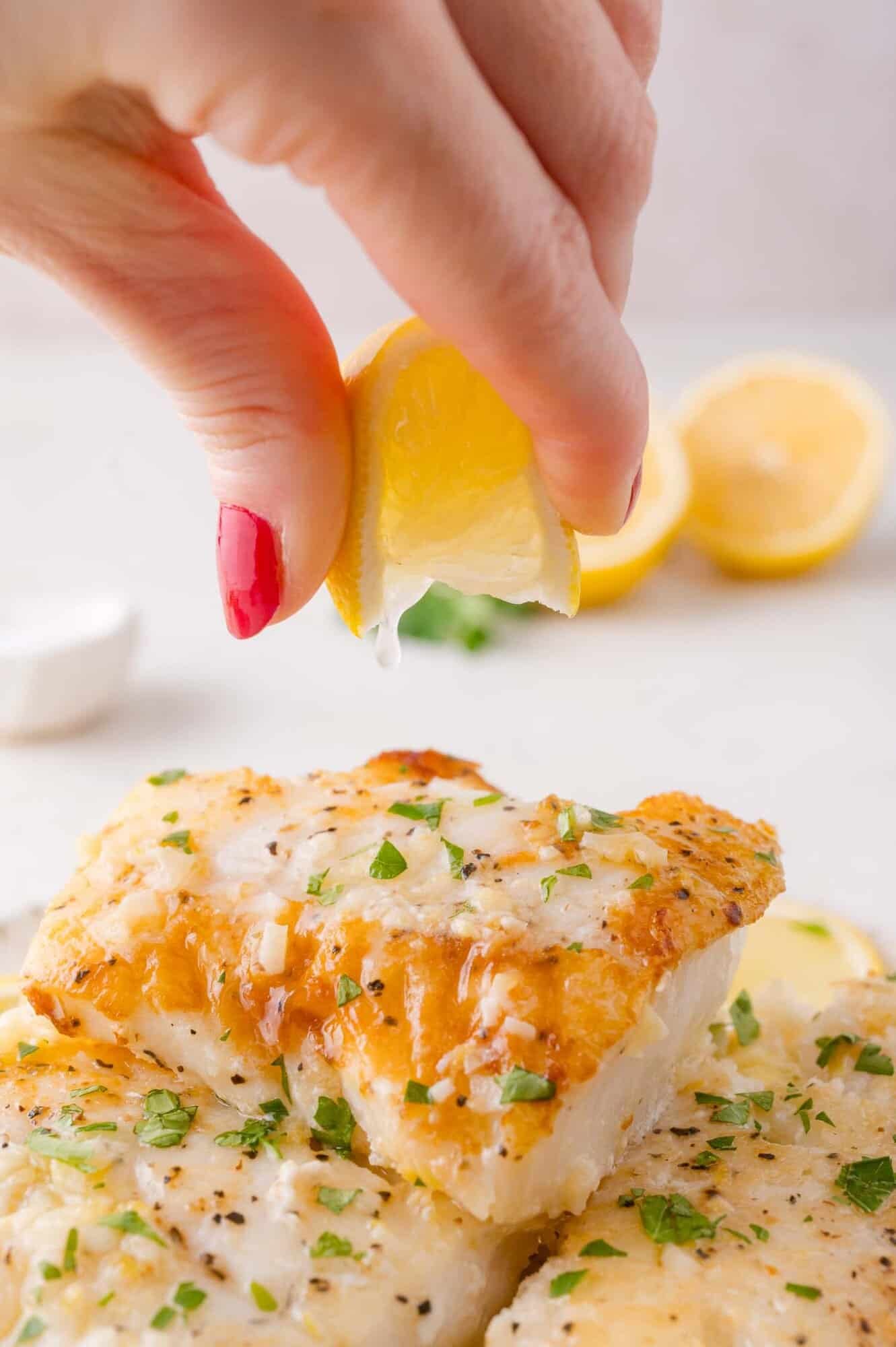 A hand with red nail polish squeezes a lemon wedge over two pieces of cooked fish garnished with chopped herbs.