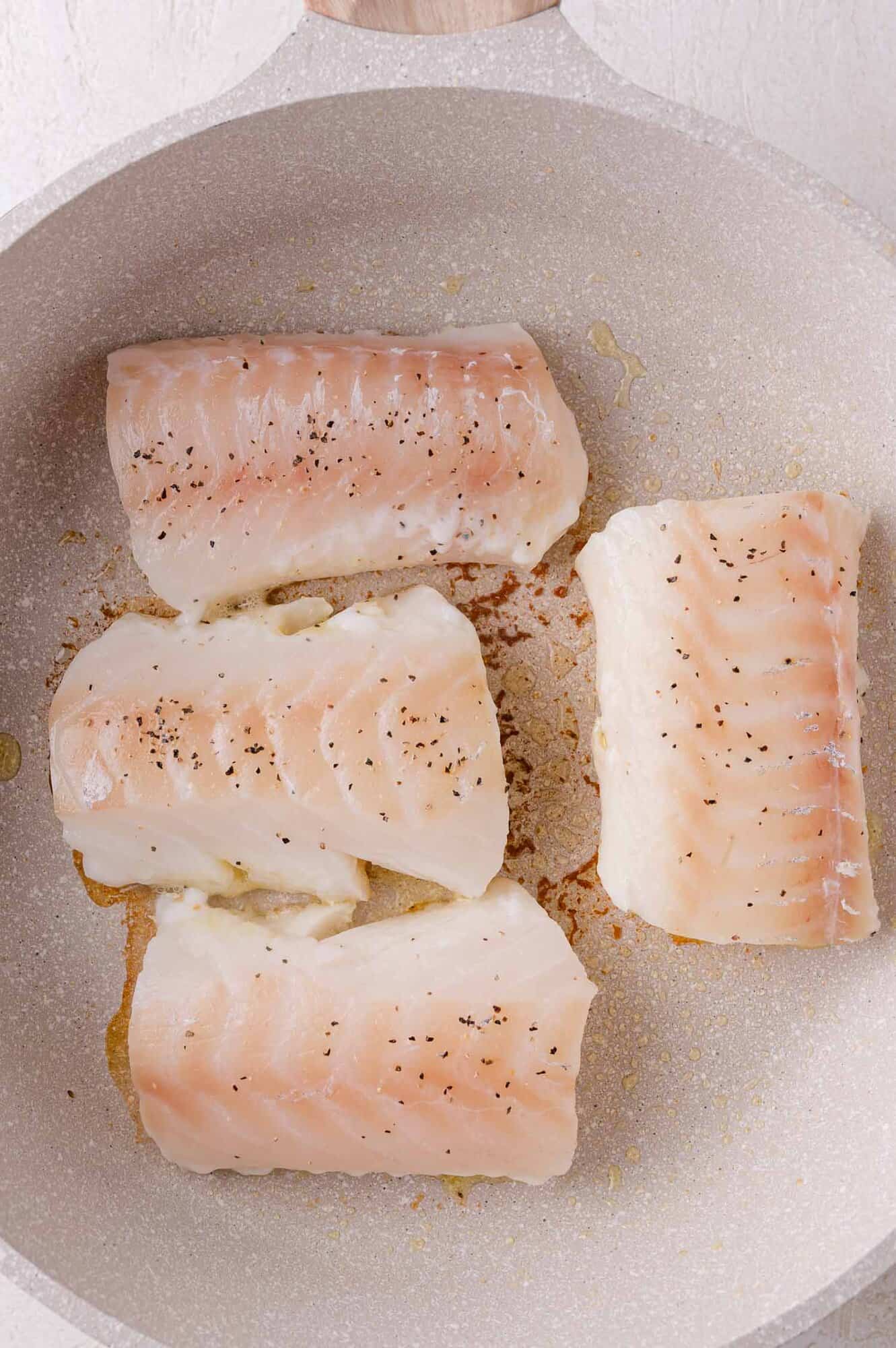 Four raw cod fillets seasoned with salt and pepper cooking in a light-colored pan.