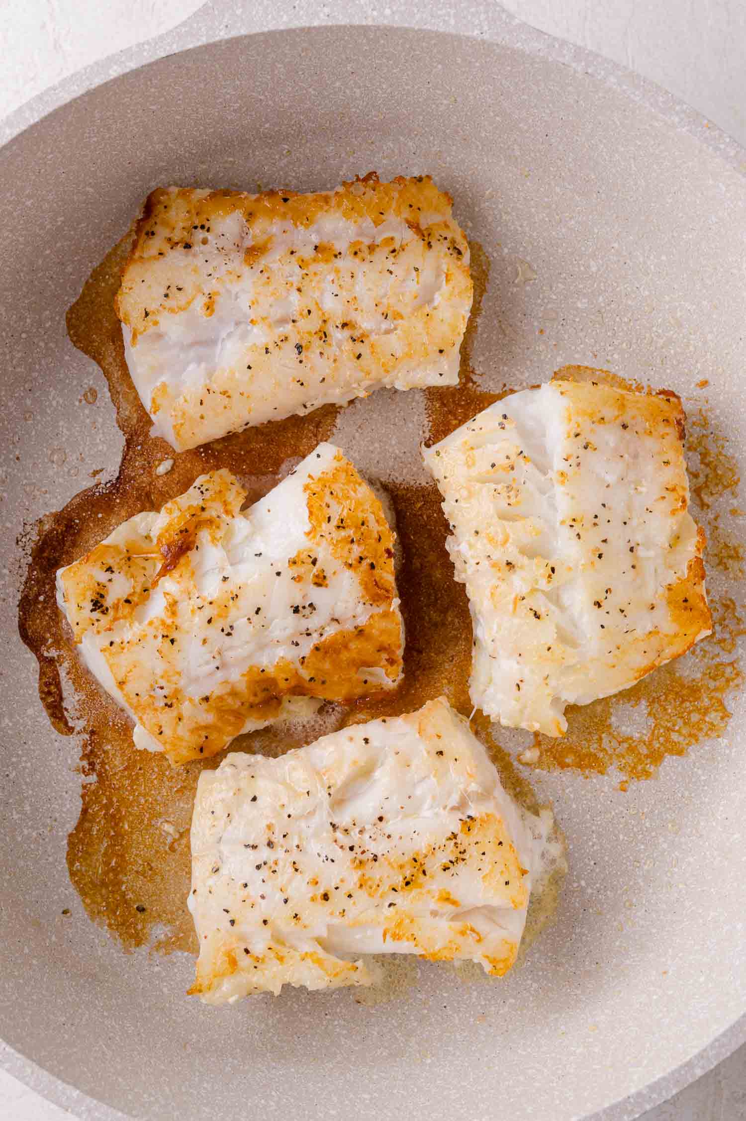 Four pieces of seasoned cod are searing in a light-colored frying pan, with golden brown edges and visible oil.