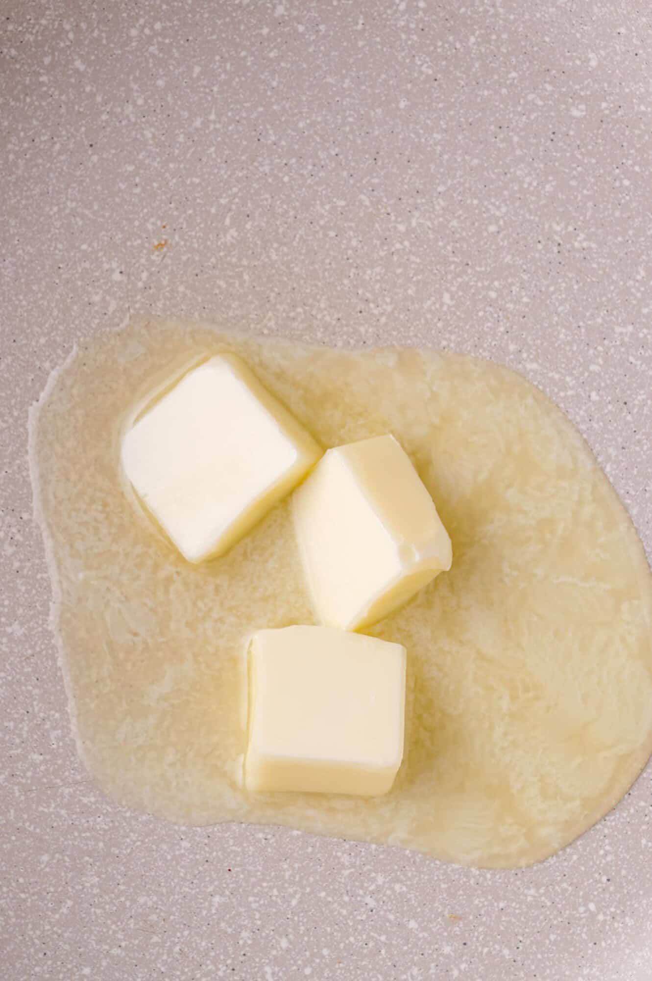 Three cubes of butter are partially melted on a light-colored pan.