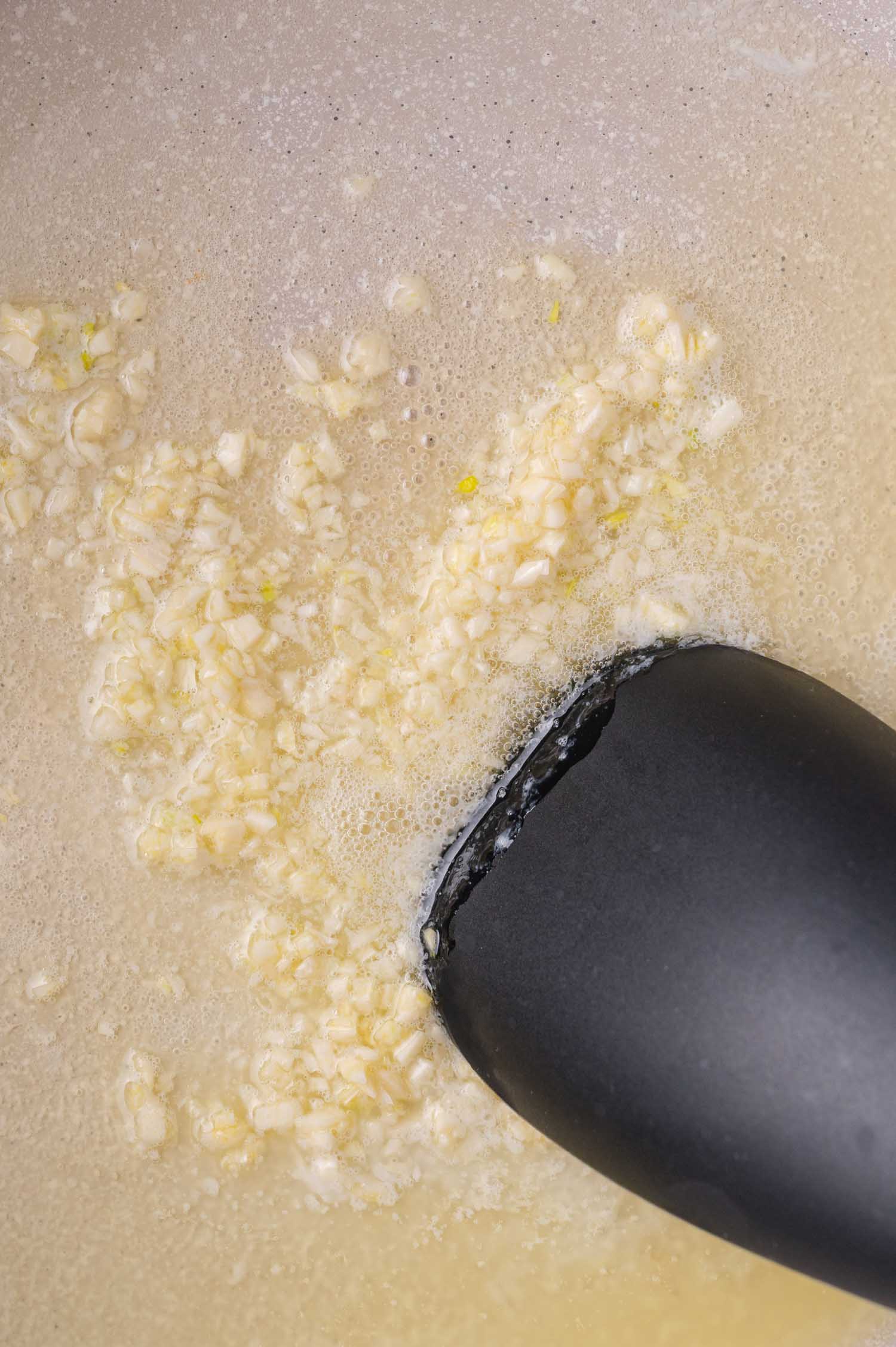 Chopped garlic being sautéed in melted butter with a black spatula in a pan.