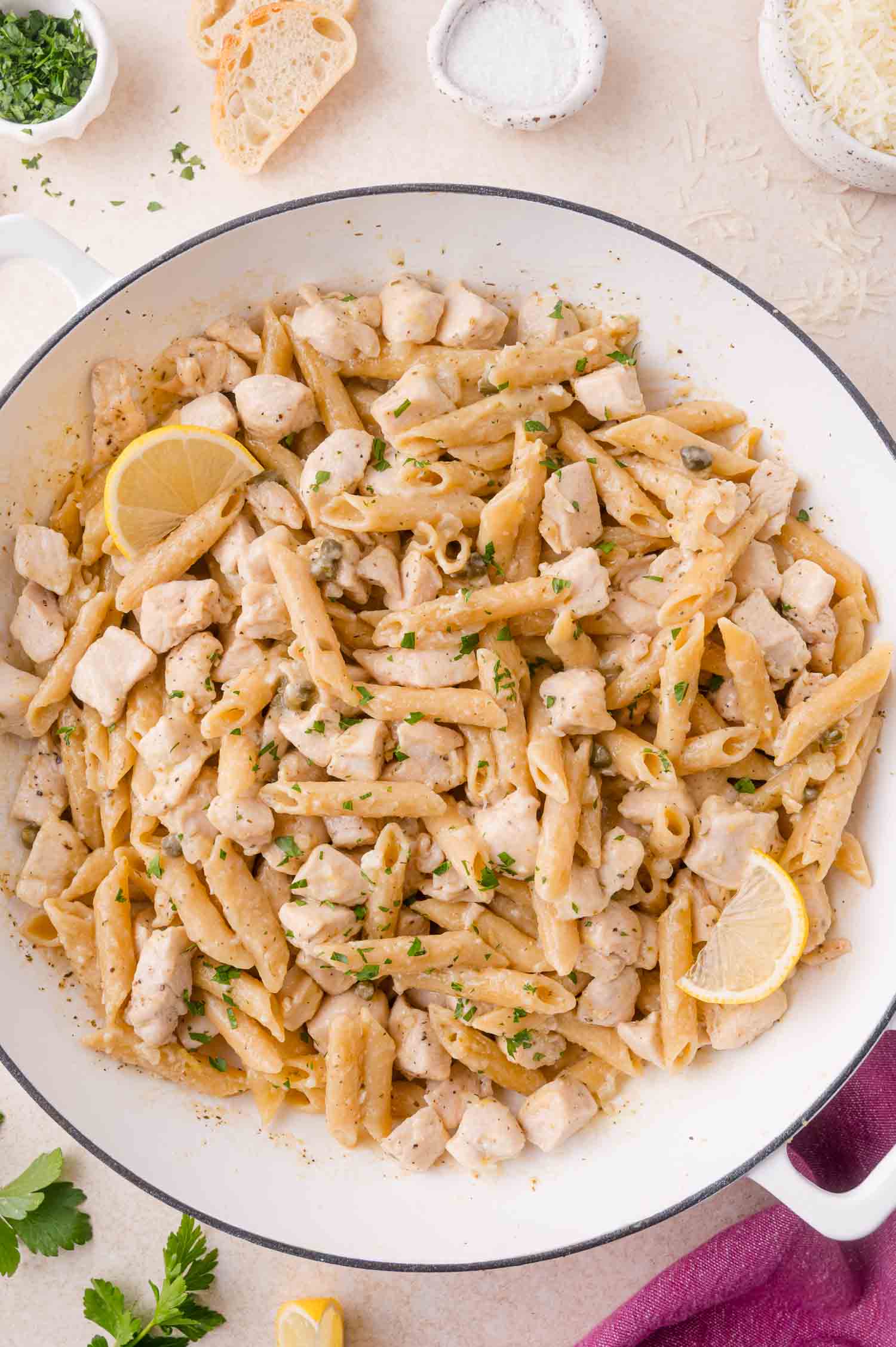 A large white pan filled with chicken piccata pasta, with cubed chicken, creamy sauce, chopped herbs, and lemon wedges. Bread slices and seasonings are visible in the background.