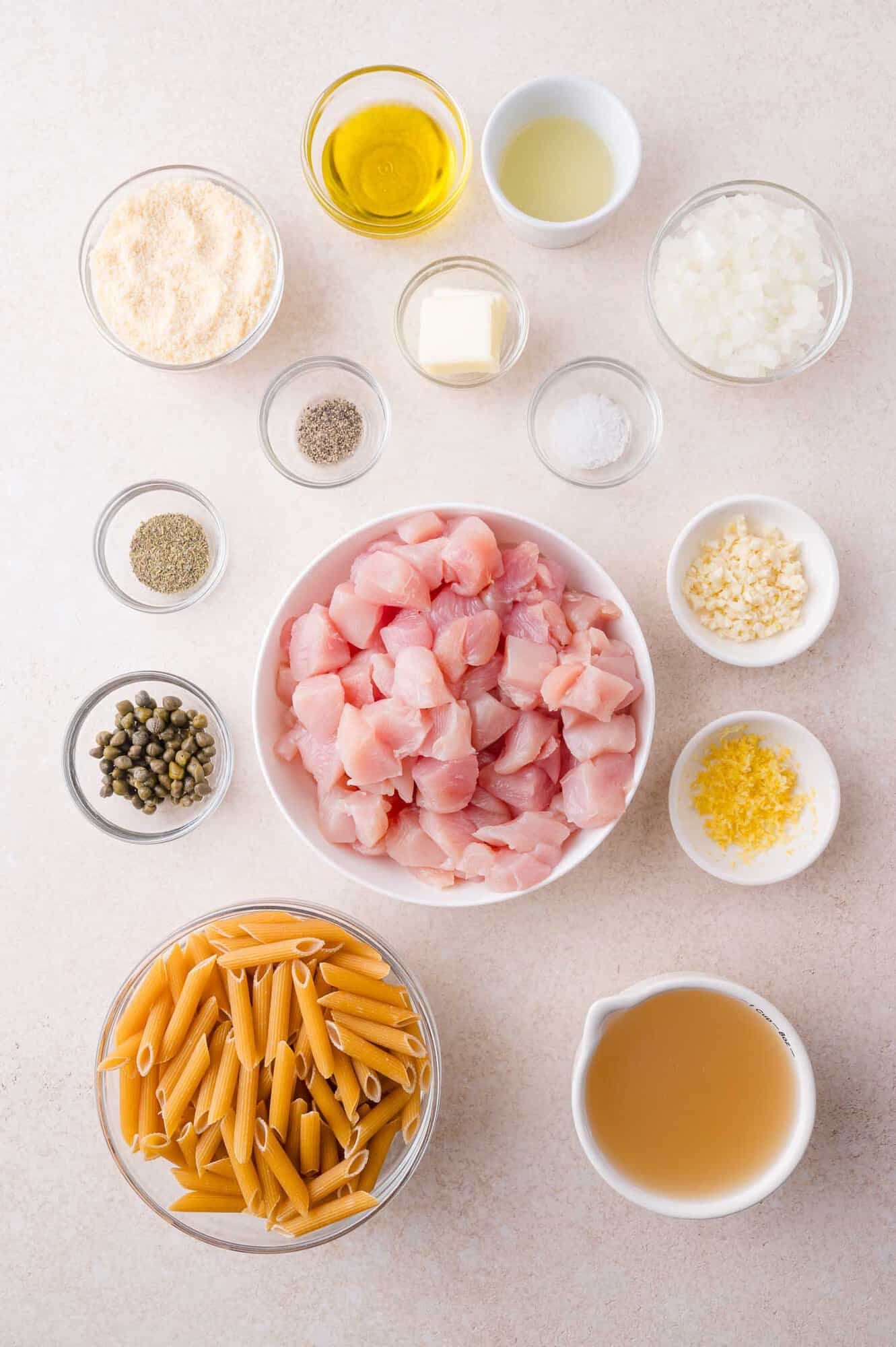 Overhead view of ingredients in small bowls on a countertop, including diced chicken, uncooked penne pasta, chopped onion, minced garlic, capers, broth, butter, oil, seasonings, and cheese.