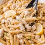 Penne chicken piccata pasta with diced chicken, lemon wedges, and herbs, mixed in a creamy sauce, served in a white dish with a black serving spoon.