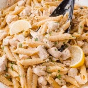 Penne chicken piccata pasta with diced chicken, lemon wedges, and herbs, mixed in a creamy sauce, served in a white dish with a black serving spoon.