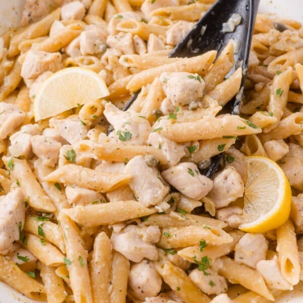 Penne chicken piccata pasta with diced chicken, lemon wedges, and herbs, mixed in a creamy sauce, served in a white dish with a black serving spoon.