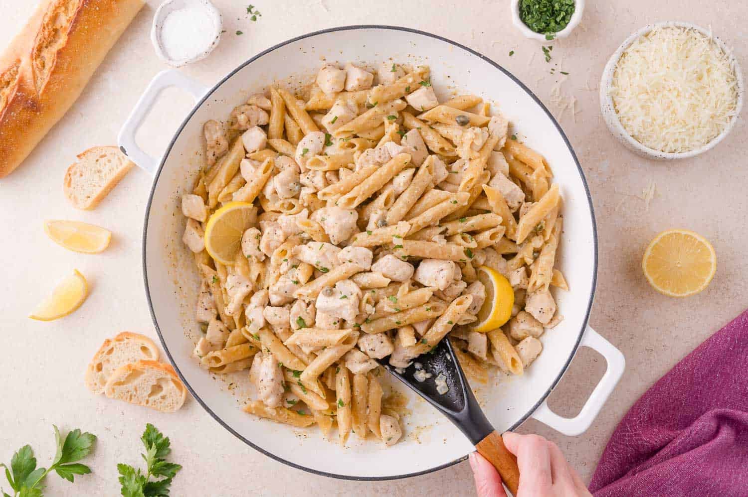 A white pan filled with penne pasta, diced chicken, lemon slices, and herbs. A hand holds a black spatula. Bread, cheese, lemon wedges, and parsley are on the table nearby.