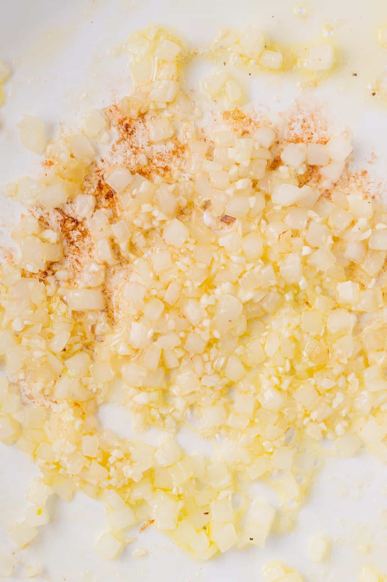 Chopped onions and minced garlic sautéing in oil with a sprinkle of spices on a white surface.