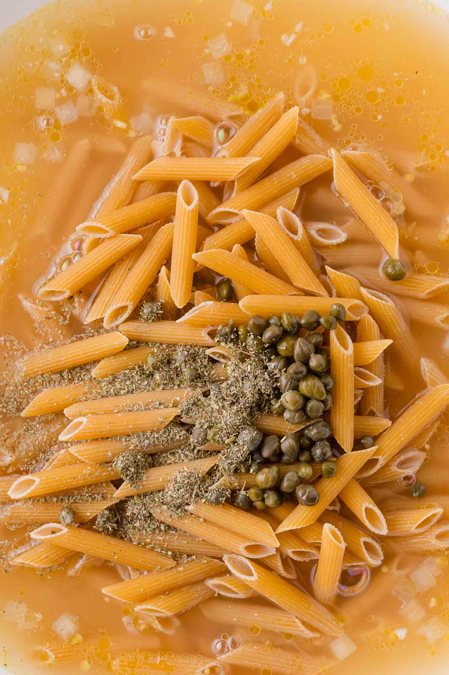 Uncooked penne pasta, capers, herbs, and spices in a broth or liquid, prepared for cooking.