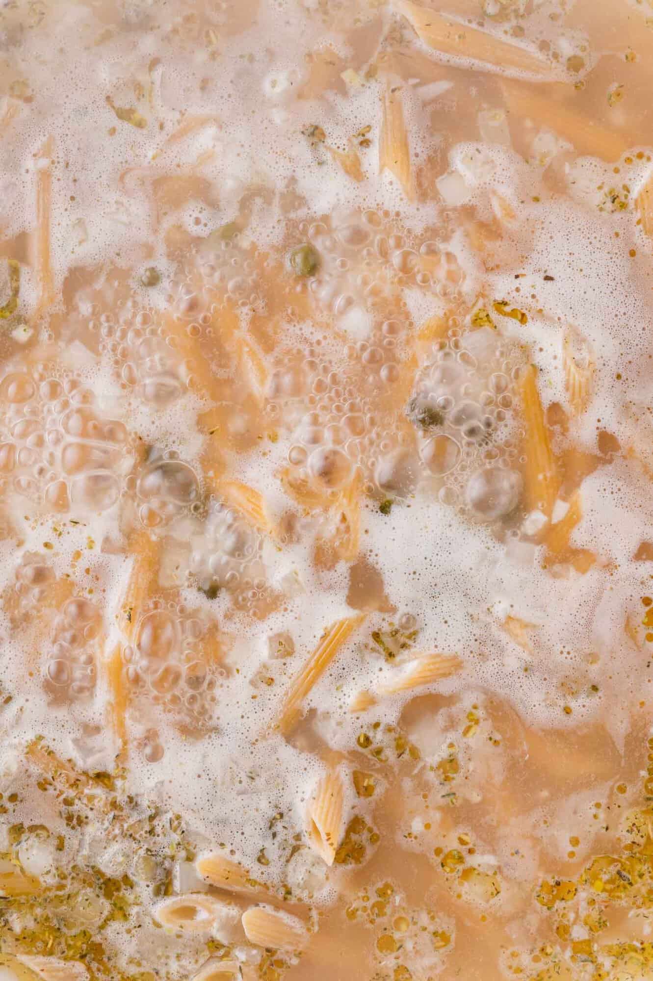 Close-up of pasta cooking in boiling water with bubbles, floating foam, and visible herbs and pasta pieces.