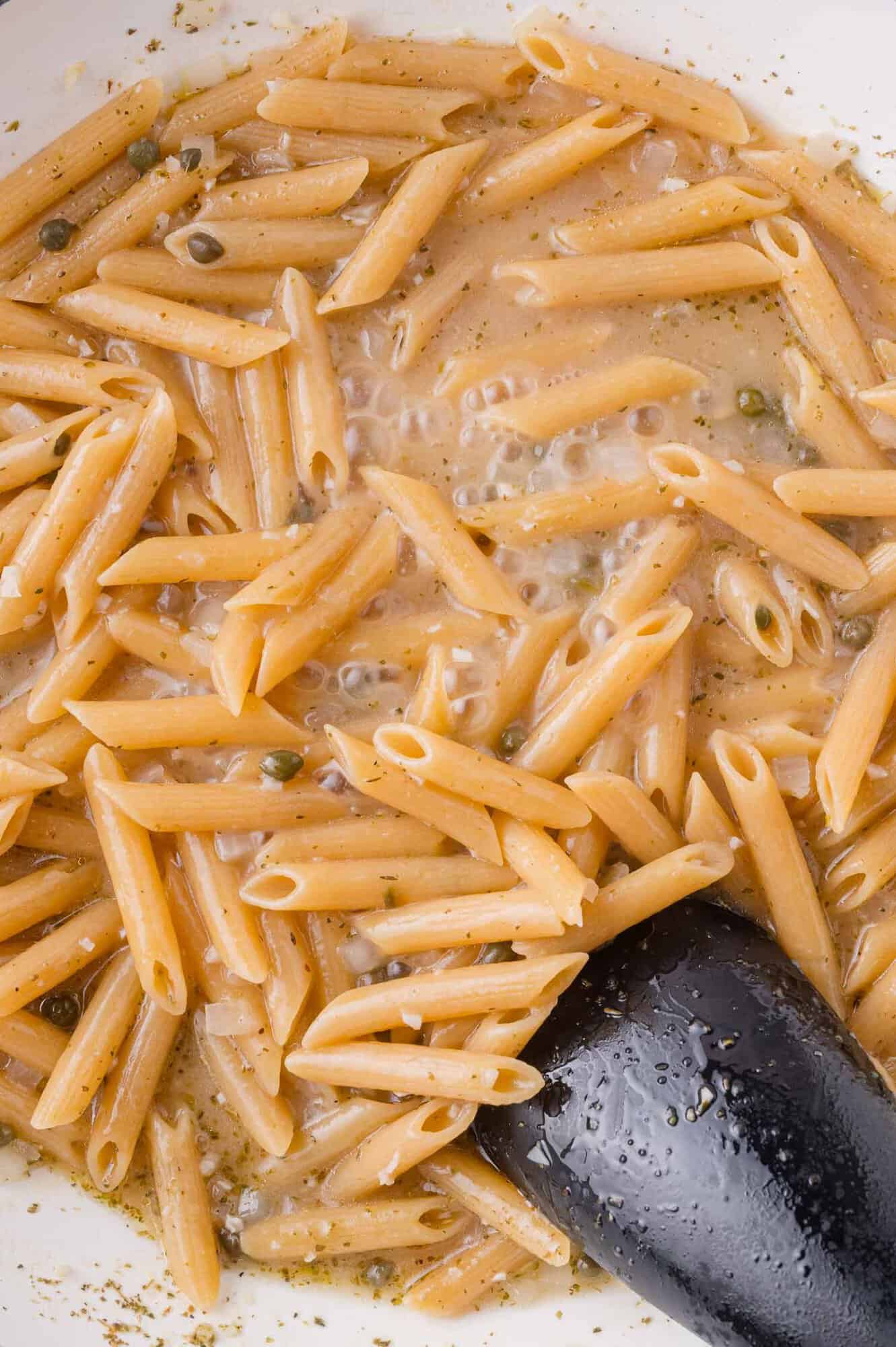 Cooked penne pasta in a creamy sauce with visible herbs and capers, stirred with a black serving spoon in a white pan.